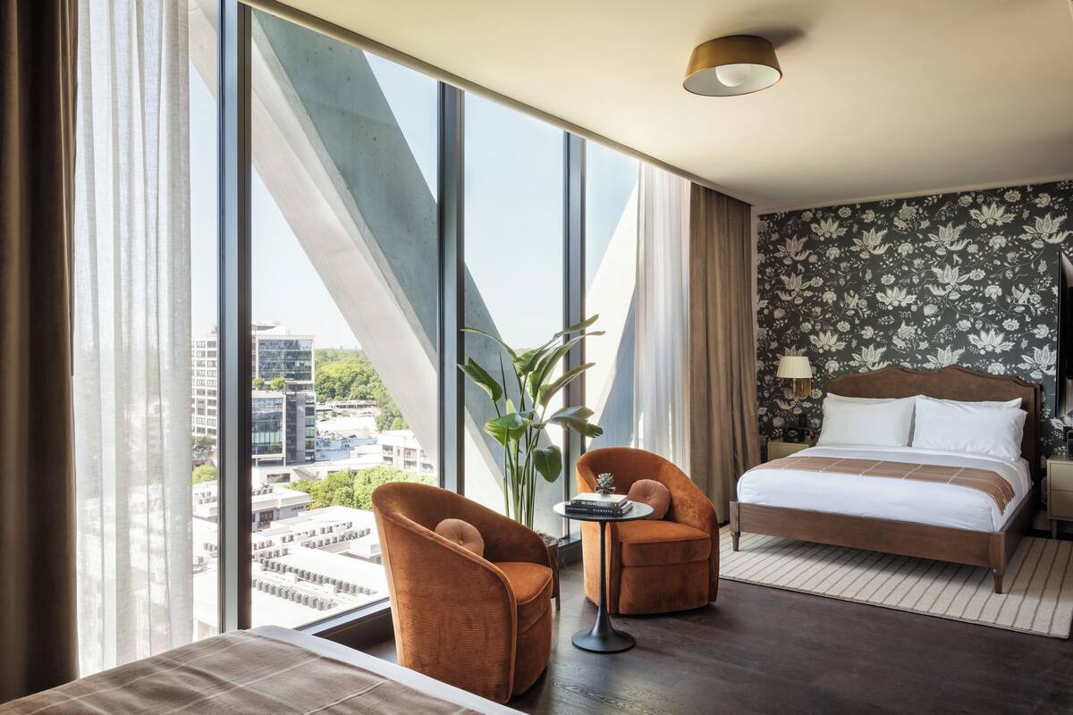 A modern hotel room with a large window, a bed, two armchairs, a small table, and a nightstand with a lamp.
