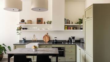 A modern kitchen with a marble countertop, a sink, a stove, and a refrigerator. There are two pendant lights, a wooden cutting board, and a bowl of fruit on the counter.
