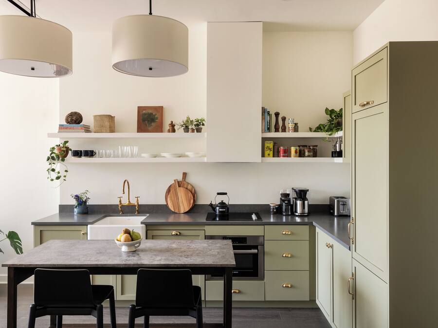 A modern kitchen with a marble countertop, a sink, a stove, and a refrigerator. There are two pendant lights, a wooden cutting board, and a bowl of fruit on the counter.