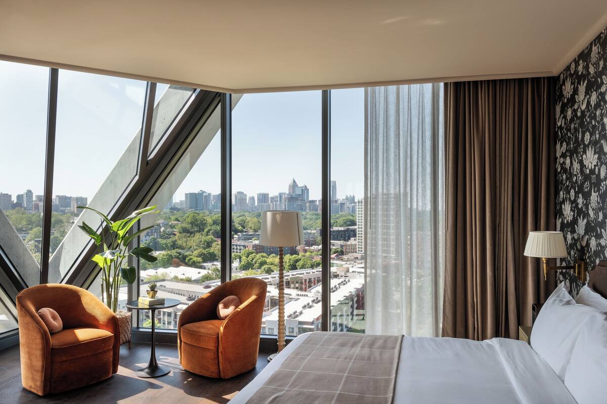 A hotel room with a large window, a bed, two armchairs, a small table, and a lamp.