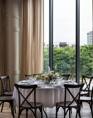 A dining area with a table set for six, large windows, and a view of greenery and buildings.