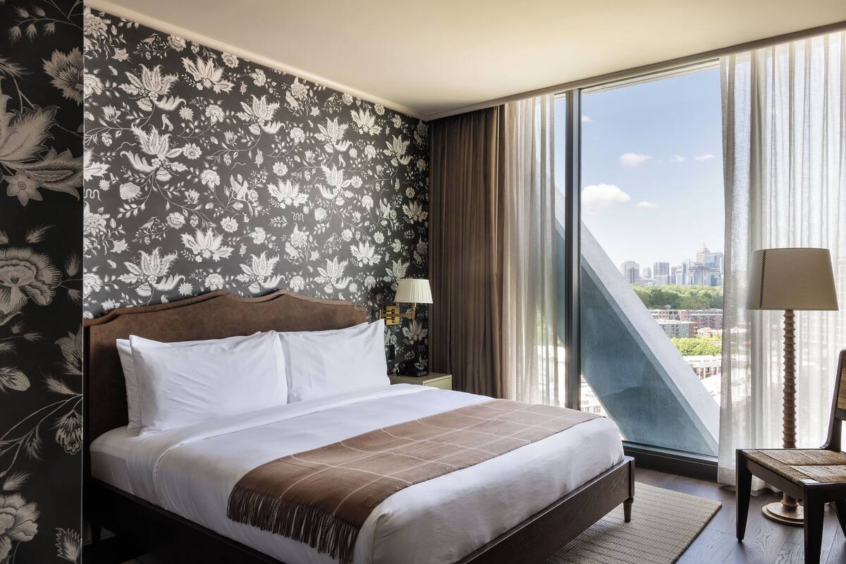 A hotel room with a large bed, a nightstand with a lamp, a chair, and a view of the city through floor-to-ceiling windows.