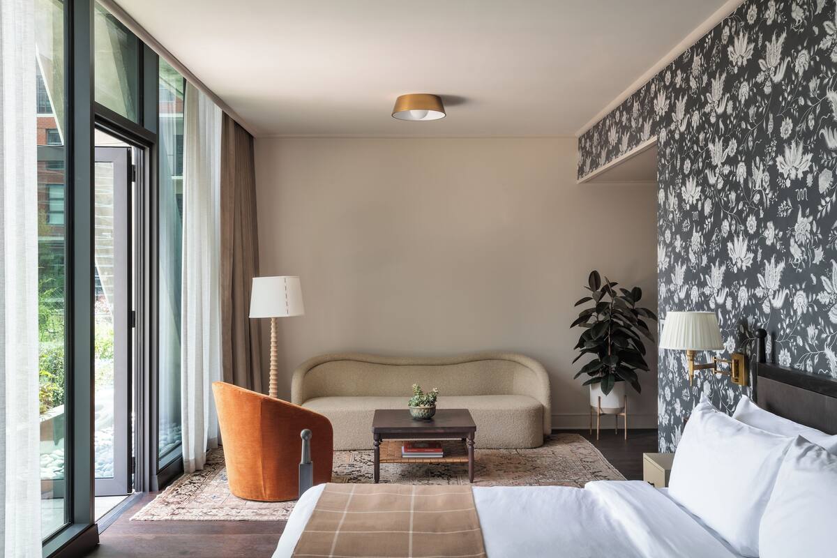 A modern hotel room with a sofa, a small table with a plant, a bed, and a large window.
