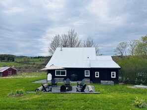 Ett svart hus med ett tak av metall, omgivet av en gräsklädd trädgård och ett rött ladugårdsbygge i bakgrunden.