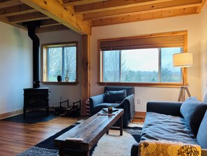 Ett mysigt vardagsrum med ett träpanelat tak, två fönster med persienner, en svart kamin, en blå soffa, ett träbord och en golvlampa.