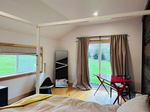 Un dormitorio moderno con cama, escritorio y silla. Hay una ventana con cortinas, un espejo y vistas a un paisaje verde.