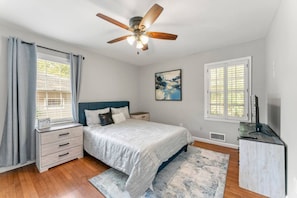 Quarto com cama, ventoinha no teto, mesa de cabeceira, cômoda e janela com cortinas.