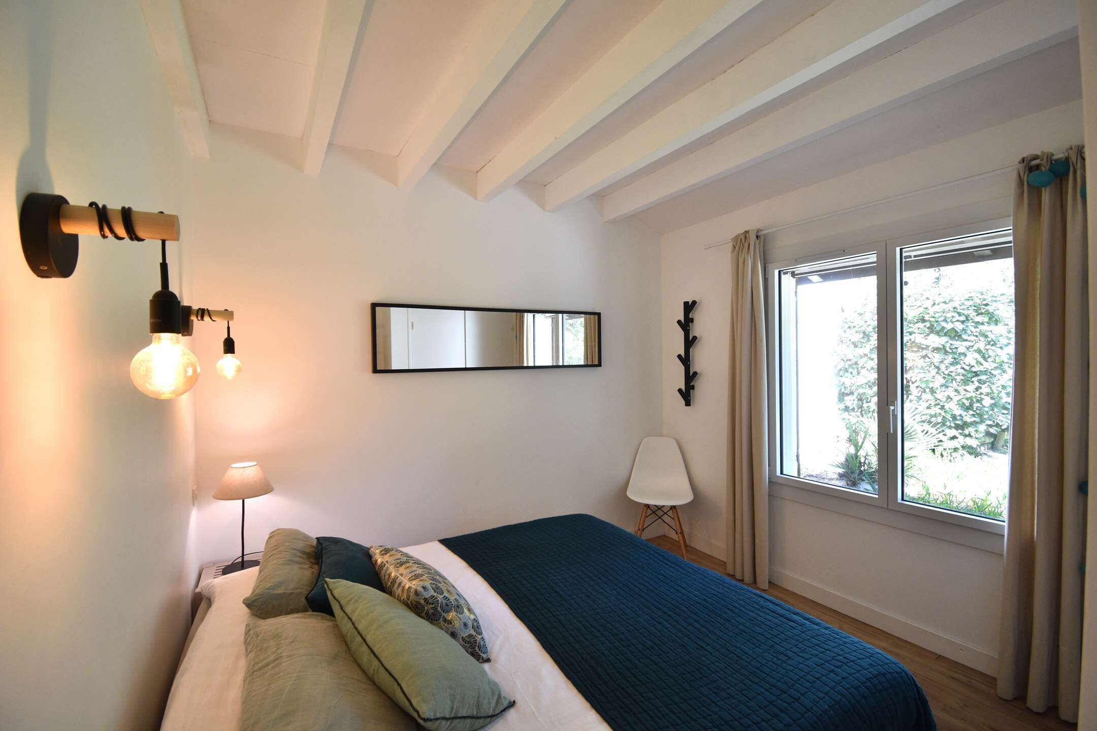 Une chambre à coucher avec un lit, une lampe de chevet, une fenêtre avec des rideaux et un miroir fixé au mur.