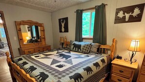 Een slaapkamer met een houten bed, een plafondventilator, een raam met gordijnen, een spiegel en een muur met decoratie van een hertenhoofd.