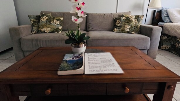 A living room with a sofa, a coffee table with a book and a potted plant, and a painting on the wall. - 407 ❤️LOCATION❤️ 5min to 🏄♀️🏊♀️🐠🦈🌸 free parking 🚙🅿️, fast WIFI💻 (Honolulu)