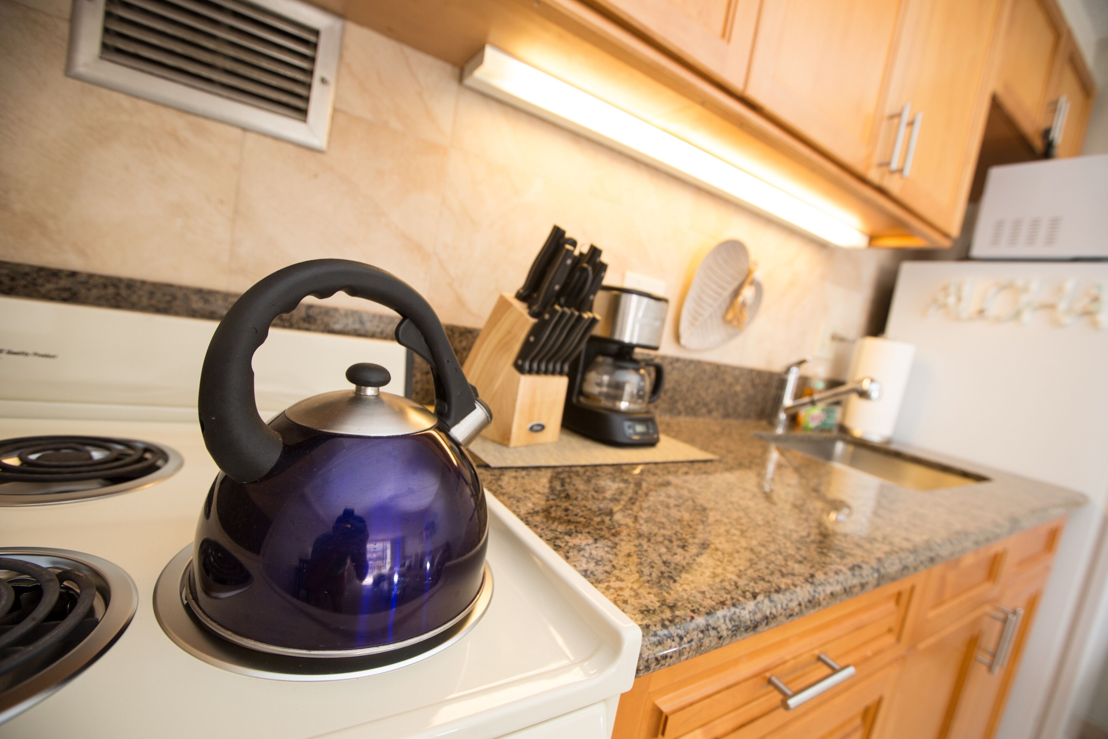 A kitchen with a stove, a kettle, a coffee maker, and wooden cabinets.