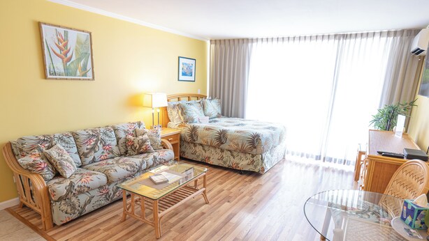 Una sala de estar con sofá, mesa de centro de vidrio, cama con ropa de cama floral, lámpara y planta.
