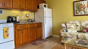 Eine kleine Küche mit Holdekor-Schränken, einem weißen Kühlschrank, einem Herd und einer Kaffeemaschine. Im angrenzenden Wohnbereich befindet sich ein Sofa mit Blumenmuster.