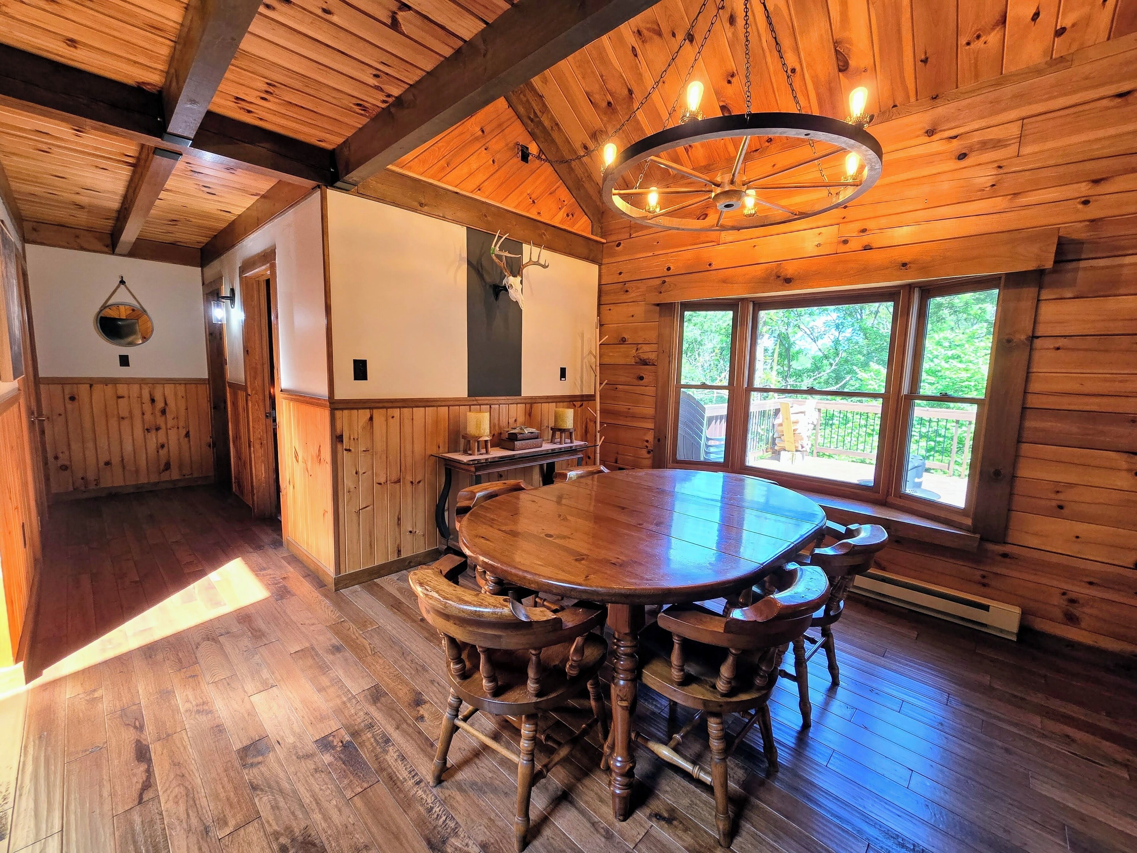 Un intérieur en bois avec une table à manger ronde, des chaises, un lustre et une grande fenêtre donnant sur un paysage verdoyant.