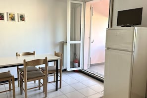 A room with a dining table, chairs, a refrigerator, a television on top, and a sliding glass door leading to an outdoor area.