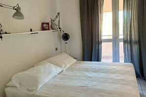 A bed with white linens, a bedside table with a lamp, a wall-mounted fan, and a window with curtains.