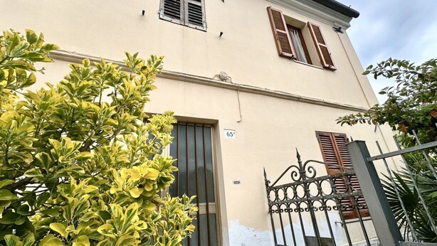 Un edificio de dos pisos con fachada color beige, una reja metálica y el número 65 en la pared.