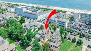 Luchtbeeld van een kustplaats met een opvallend hotel, een ruime parkeerplaats, een strand op de achtergrond en een rode pijl die naar het hotel wijst.