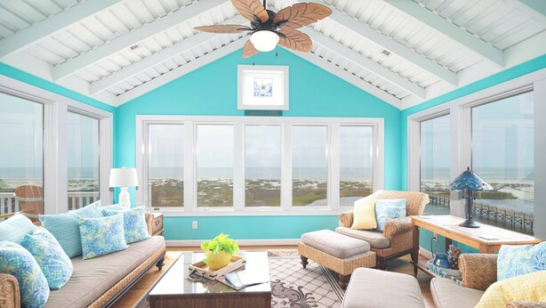 A sunroom with a view of the ocean, furnished with wicker furniture and a ceiling fan. - Hobbs Realty - "All that Jazz" includes a private pool & panoramic views! (Holden Beach)