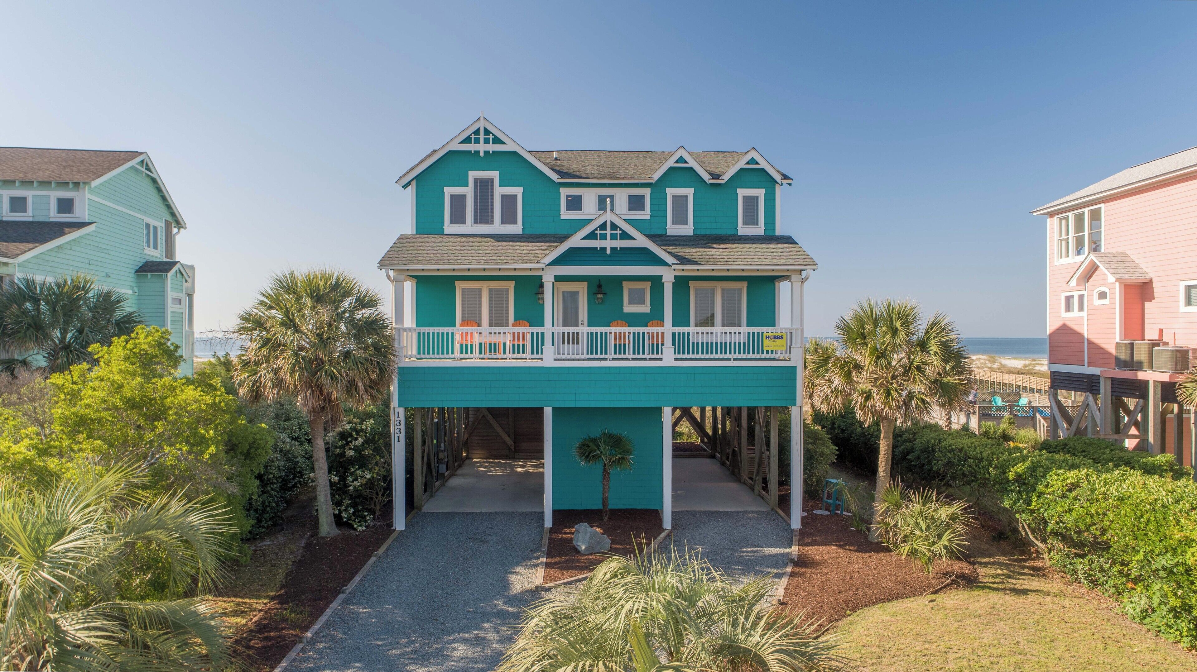 A two-story house with a balcony, surrounded by palm trees and a clear view of the beach.