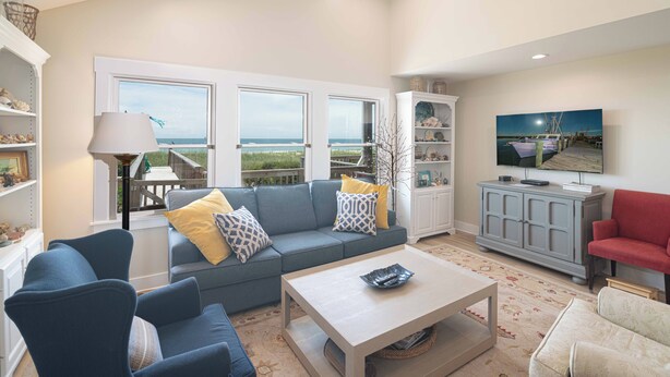 A living room with a sofa, armchairs, a coffee table, and a television. There are large windows with a view of the outdoors. - Hobbs Realty - "Fore Reel" an oceanfront dog friendly home that offers amazing views! (Holden Beach)