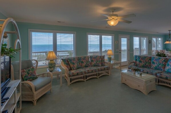 A spacious living room with wicker furniture, large windows, and a ceiling fan.
