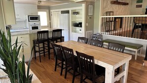 A dining area with a wooden table and chairs, a kitchen in the background, and a staircase leading to a balcony.