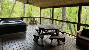 A wooden deck with a hot tub, picnic table, and benches, overlooking a forested area.