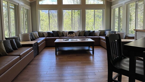 A spacious room with a large sectional sofa, a wooden coffee table, and a dining table. The room features a vaulted ceiling and large windows offering a view of the outdoors.