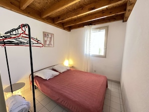 Una cama individual con una colcha roja estampada, un tendedero de pared, una mesita con una lámpara y una ventana con cortina.