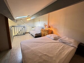 A bedroom with a bed, a chair, a nightstand, a lamp, a fan, and a staircase.