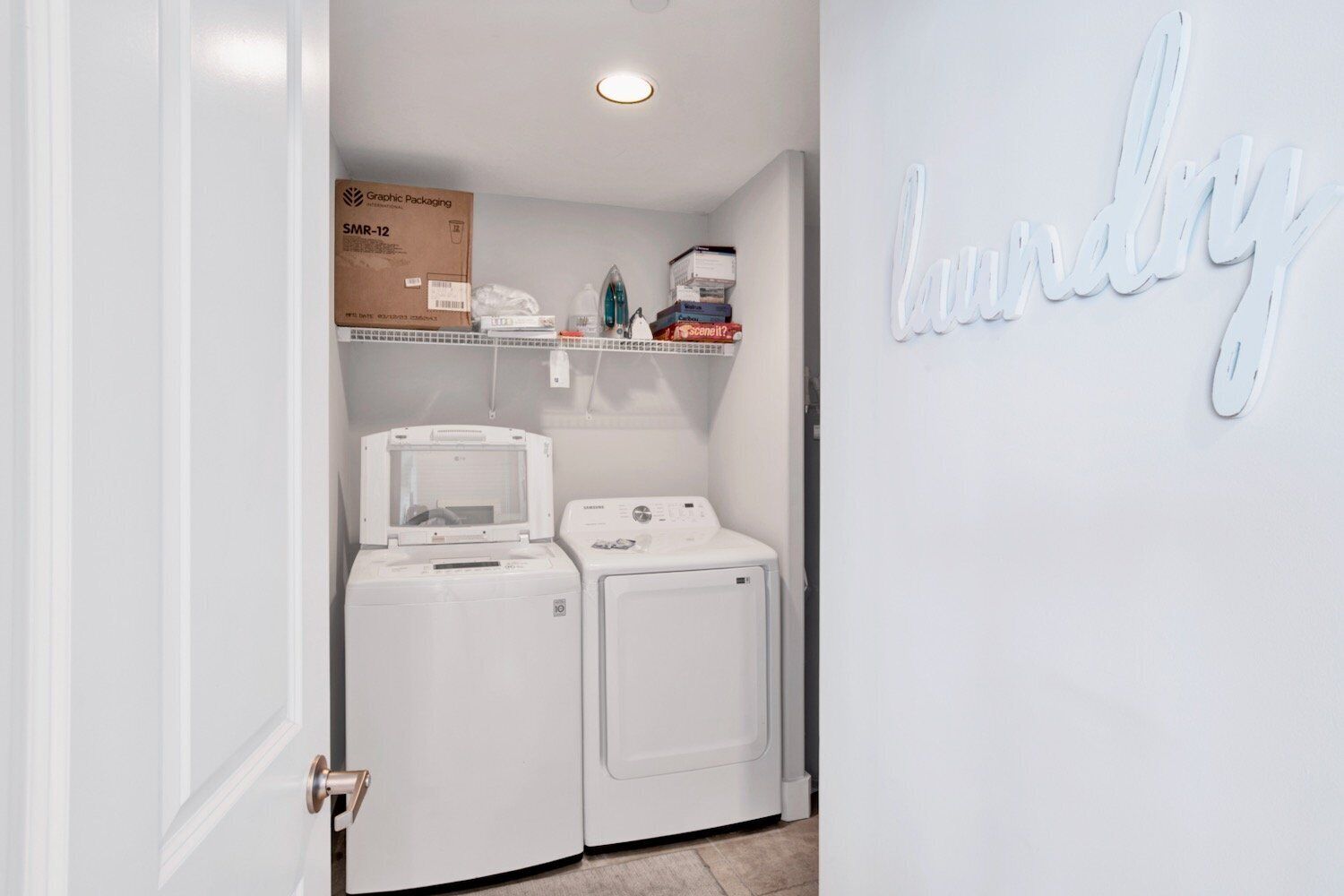 A laundry room with two white washing machines, a shelf with books and a box labeled 'Graphic Packaging SMR-12'.
