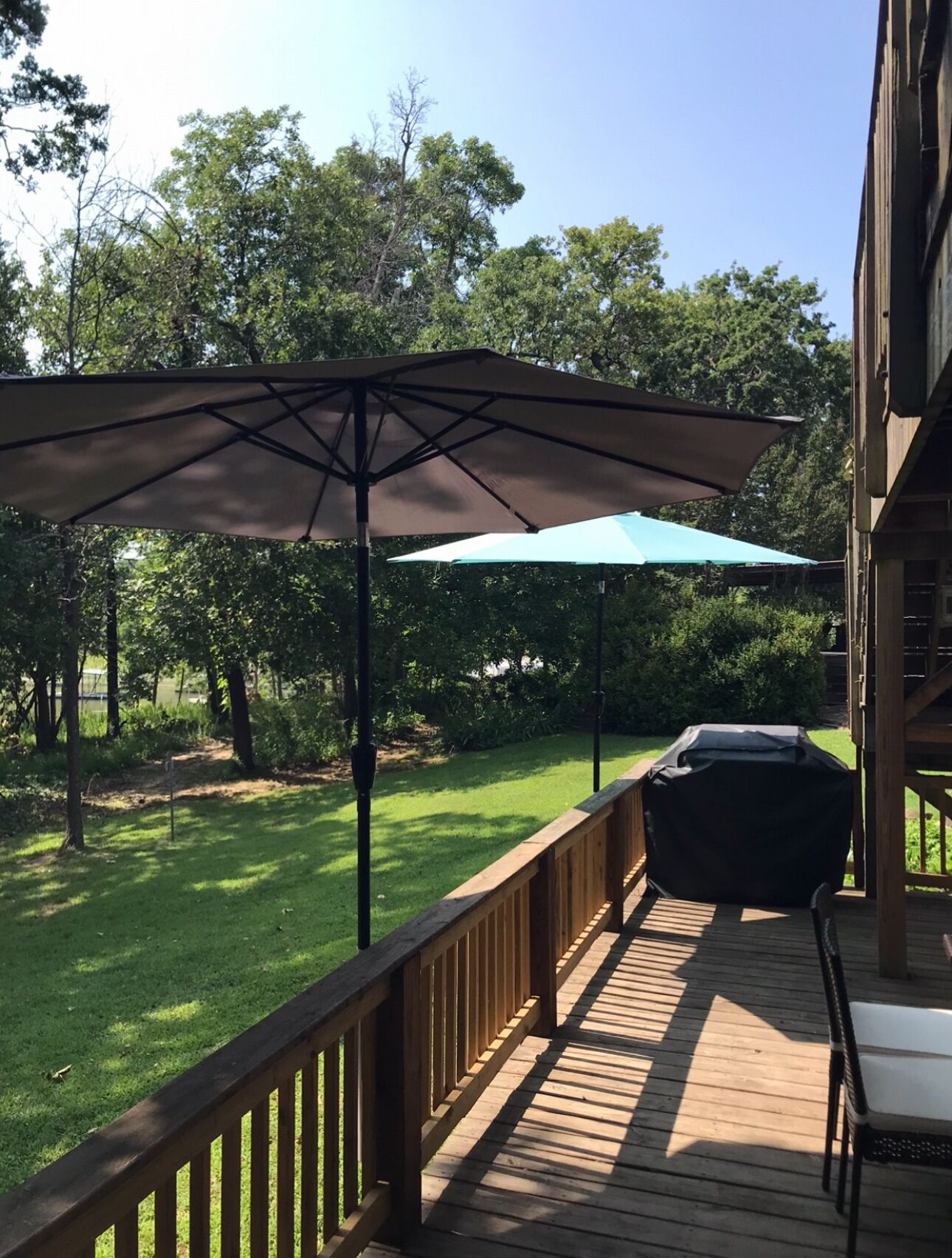A wooden deck with a barbecue grill and two umbrellas, surrounded by a grassy yard and trees.