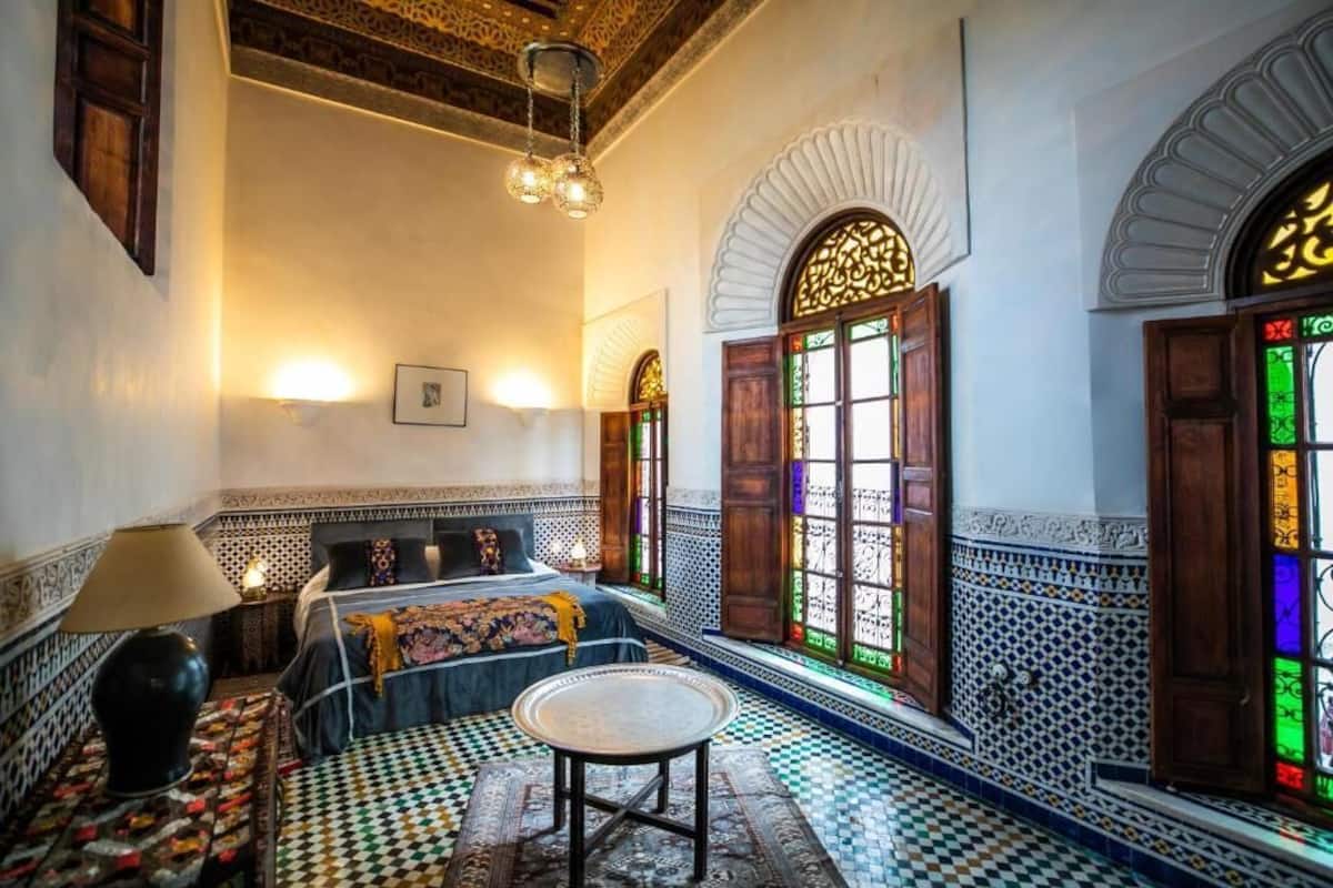 Una habitación con piso a cuadros, una cama con colcha estampada, una mesa redonda y una lámpara de pared.