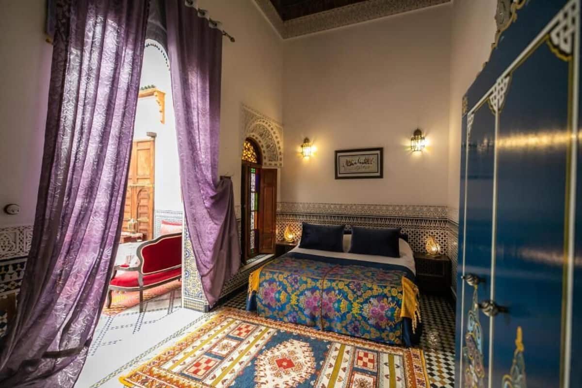 Un dormitorio de estilo marroquí con una colcha estampada, una silla roja y una alfombra decorativa.