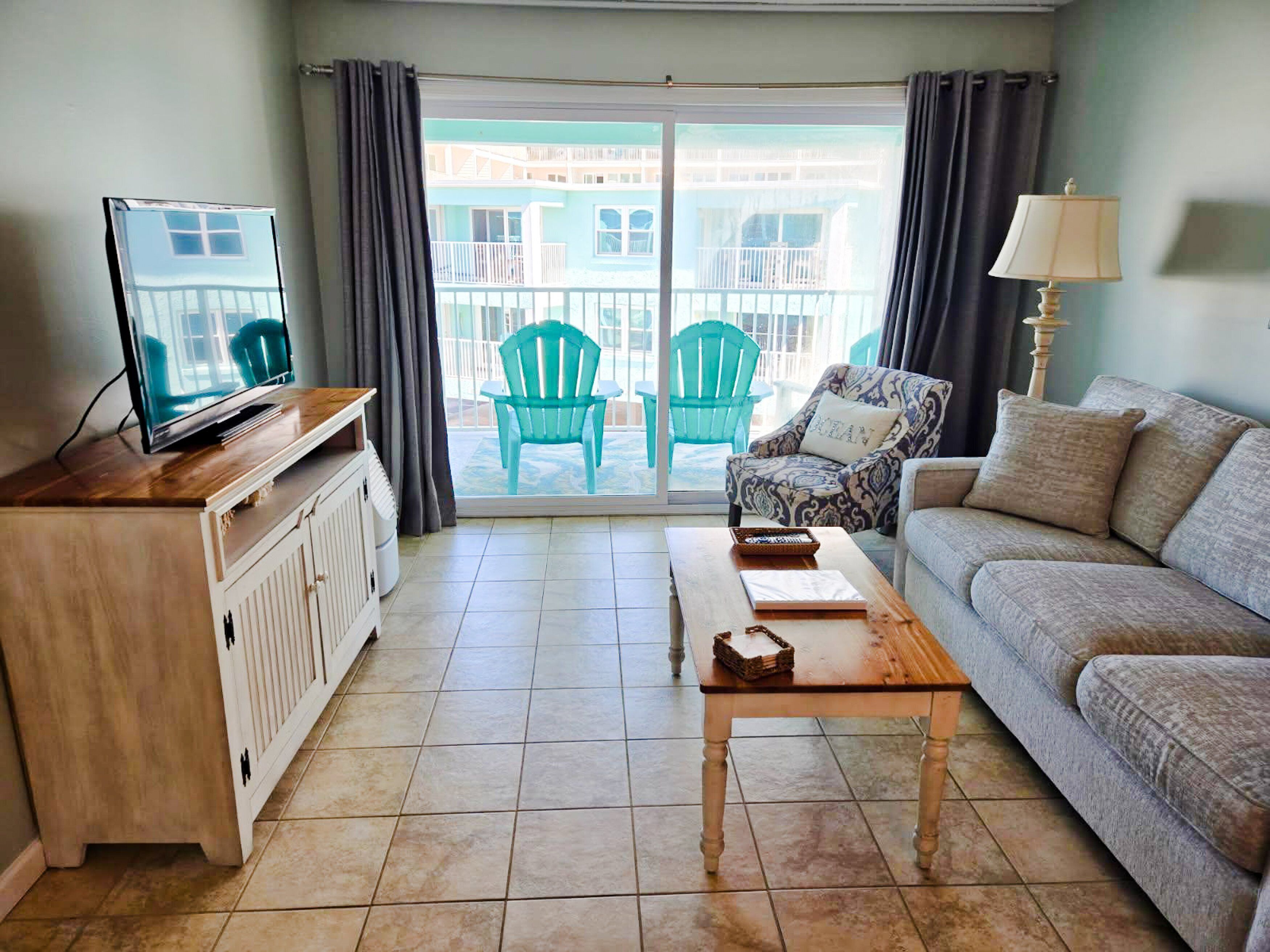 Een woonkamer met een televisie, een bank, een bijzettafeltje en uitzicht op een balkon met stoelen.