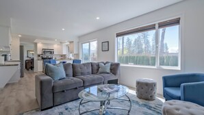 A modern living room with a grey sofa, a glass coffee table, and a view of the kitchen and outdoors.