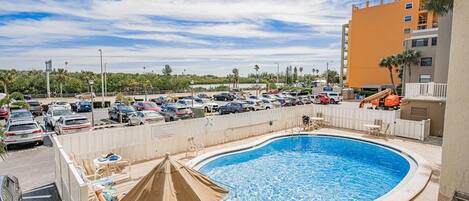 Una piscina con sillas de descanso y sombrillas, rodeada por un estacionamiento y un edificio.