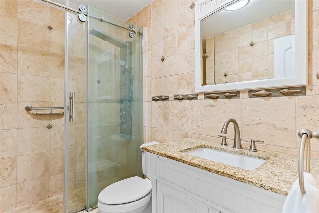 Een badkamer met een douchecabine van glas, een wit toilet, een wastafel met een granieten blad en een spiegel boven de wastafel.