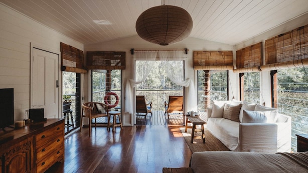 A spacious living room with a wooden floor, a white sofa, and a large window offering a view of the outdoors. - Boathouse 2 - Hunter Valley Couples Retreat, Bathhouse 2 at Leaves and Fishes (Lovedale)