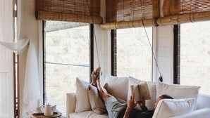 A person relaxing on a white sofa with a book, a wooden table, and a floor lamp in a room with large windows and bamboo blinds. - Boathouse 2 - Hunter Valley Couples Retreat, Bathhouse 2 at Leaves and Fishes (Lovedale)