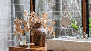 Un baño con tocador de madera, un espejo redondo y un ventanal con vistas a plantas tropicales.