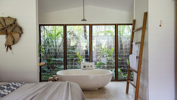 Un baño con una bañera independiente, una escalera de madera y un ventanal con vistas a la vegetación.