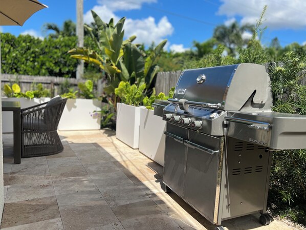 Een terras buiten met een barbecue van roestvrij staal, een rieten stoel en potplanten.