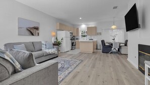 En moderne stue med en grå sofa, et spiseområde med bord og stole, et køkken med træskabe, og et fjernsyn monteret på væggen.
