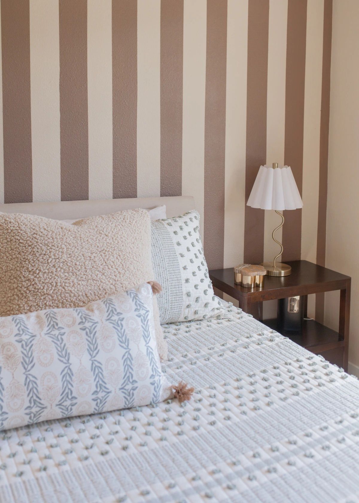 Un lit recouvert d’une couverture blanche à motifs, deux oreillers, une table de chevet avec une lampe et un mur à rayures.