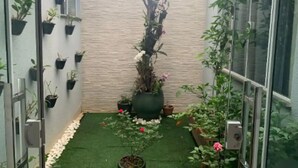 Ein schmaler Außenbereich mit verschiedenen Kübelpflanzen und Blumen, eine kleine Gartenanlage mit Kunstgrashalm und eine Backsteinmauer.