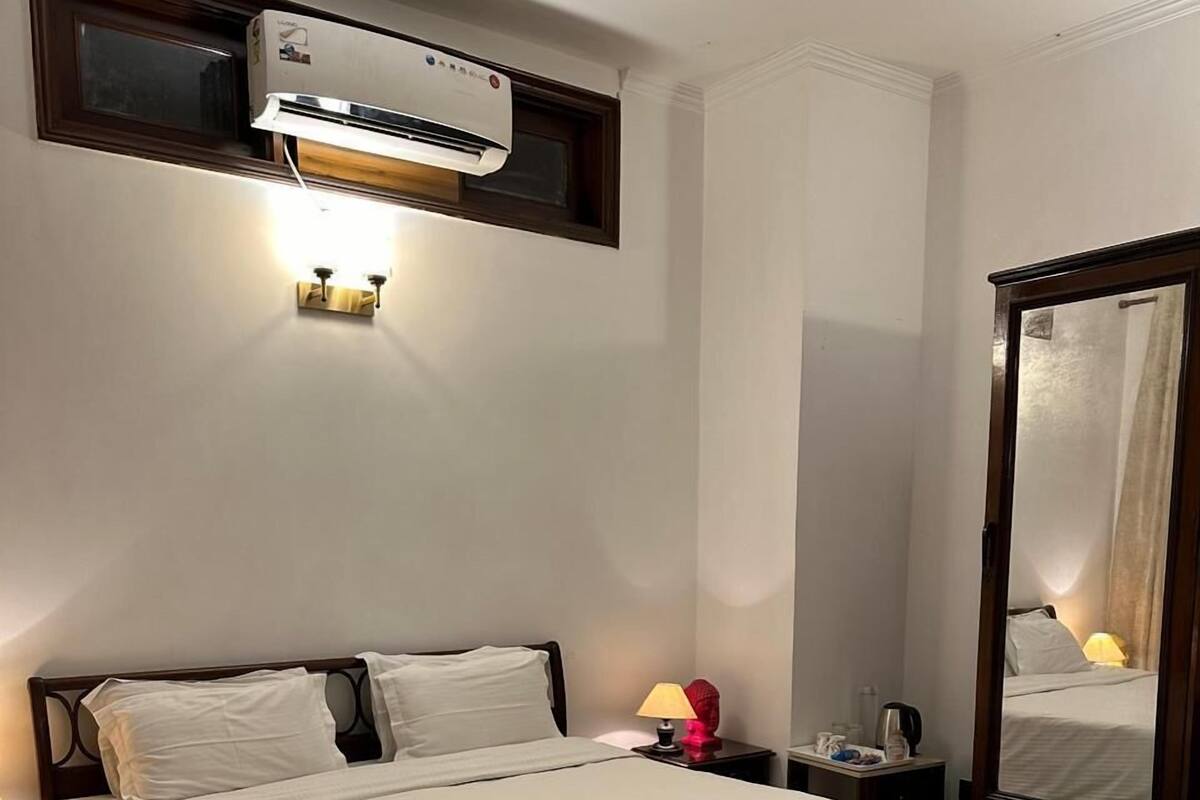 Una cama bien hecha en una habitación de hotel, con un espejo grande, una mesita de noche con lámpara y un equipo de aire acondicionado en la pared.
