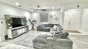 A living room with a sectional sofa, a flat-screen TV, a ceiling fan, and a potted plant. - New-North Port Florida -Heated pool - 5 BED (North Port)