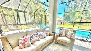 A sunlit outdoor seating area with a wicker sofa and armchairs, a small table, and a view of a swimming pool. - New-North Port Florida -Heated pool - 5 BED (North Port)